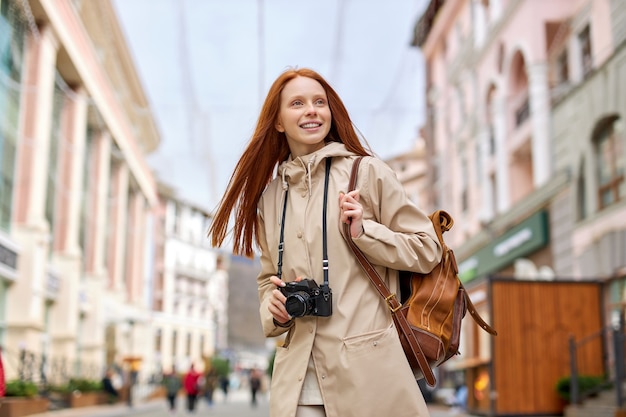 Счастливая женщина в бежевом пальто фотографирует с ретро пленочной камерой, портрет