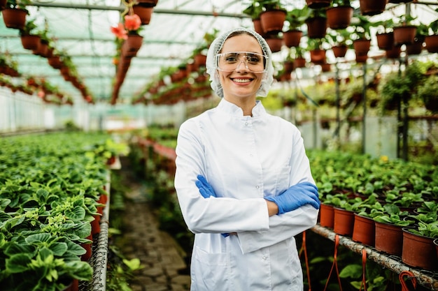 Happy female biologist with crossed arms standing at plant nursery and looking at camera