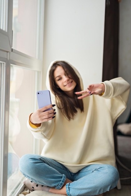 ソーシャルメディアの投稿のためにスマートフォンのカメラで自分撮りの肖像画を撮る幸せな女性ブロガー携帯電話を介して家族や友人とビデオ通話で話している陽気な10代の少女ソフトセレクティブフォーカス