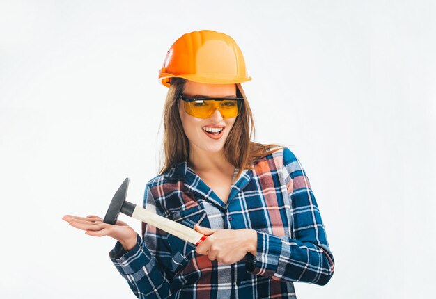 Happy female in building helmet with hammer and orange protective glasses isolated on white background Young model wearing helmet and glasses