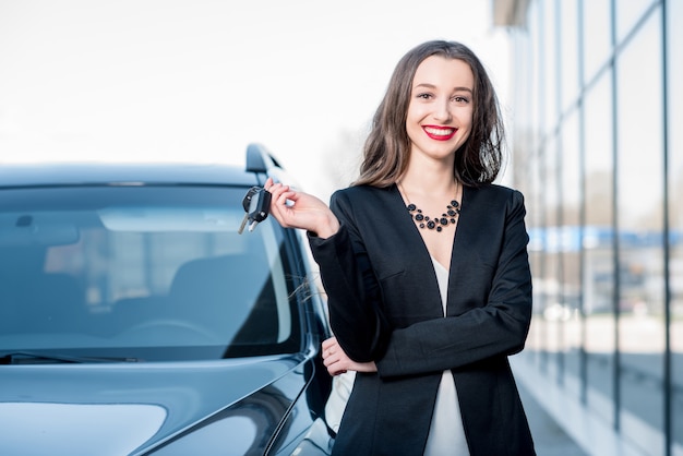 Happy female buyer holding keys near the car in front of the modern avtosalon building