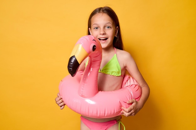 Happy female child playing with inflatable flamingo ring enjoy summer at tropical beach resort creating joyful memories of her childhood holidays standing isolated over yellow background