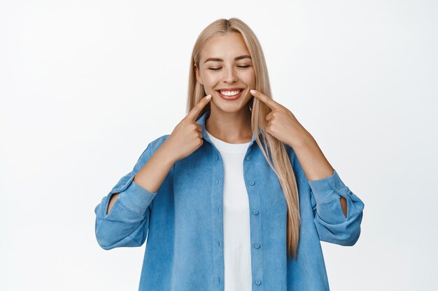 Happy female client showing her whitened healthy smile, pointing fingers at teeth with satisfied face expression, standing on studio