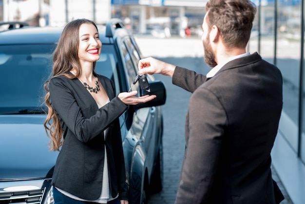 現代のavtosalonの近くに立っている新しい車から鍵を受け取る幸せな女性の顧客