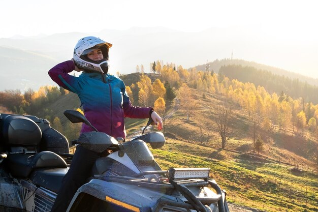 日没時の秋の山でATVクワッドバイクに乗ってオフロードを楽しんでいる保護ヘルメットの幸せな女性ドライバー。