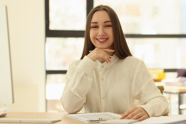 Happy female employee sits at table near papers in office smiling and looking in camera