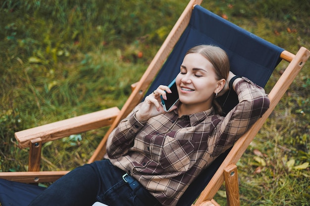 デジタルネットブックに取り組んでいる幸せな女性フリーランサーは、庭の椅子でリラックスし、リモートワーク中に公園でポーズをとるラップトップを持つカメラの女性に微笑んでいます