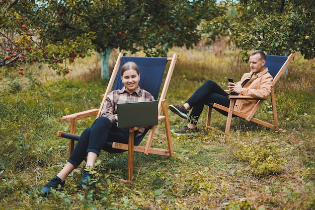 Happy female freelancer working on digital netbook relaxing in garden chair and smiling at camera woman with laptop posing in park during remote work