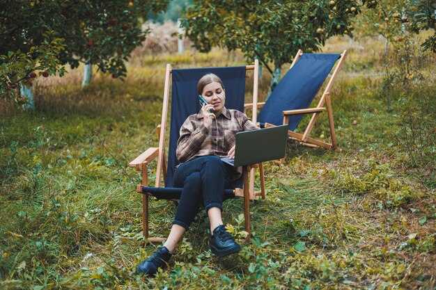 Happy female freelancer working on digital netbook relaxing in garden chair and smiling at camera woman with laptop posing in park during remote work