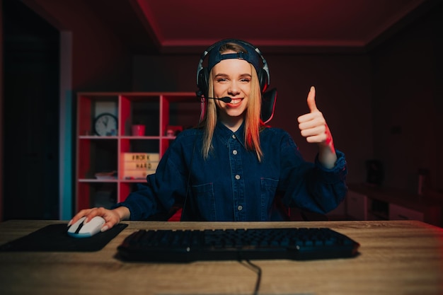 Foto giocatore femminile felice in cuffia seduto alla scrivania del computer a casa con un sorriso sul viso che guarda l'obbiettivo
