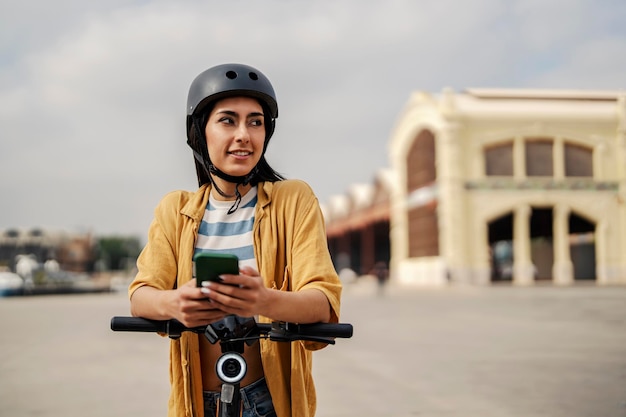 A happy female hipster is using a mobile app for renting an electronic scooter