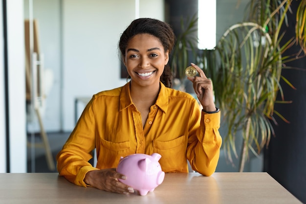 Happy female manager putting bitcoin coin in piggy bank in office interior woman smiling at camera sitting at desk