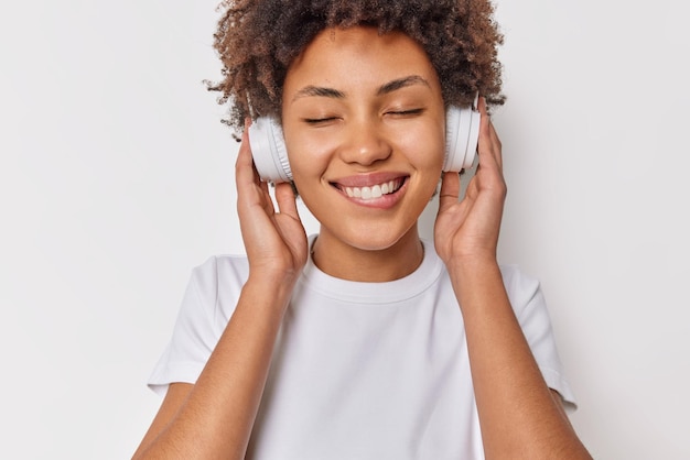 Happy female meloman enjoys sound in new headphones keeps eyes closed bites lips listens favorite music dressed casually isolated over white background. People hobby and modern technologies.