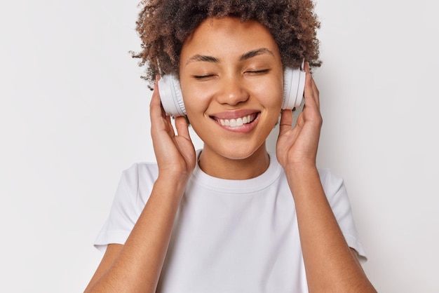 Happy female meloman enjoys sound in new headphones keeps eyes
closed bites lips listens favorite music dressed casually isolated
over white background. people hobby and modern technologies.