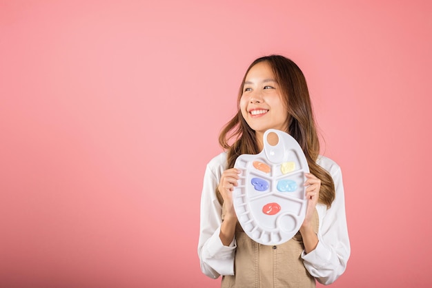 Happy female painting using paintbrush and palette with colors, Asian beautiful young woman artist holding brush and paint palette, studio shot isolated on pink background, Paintings and art equipment