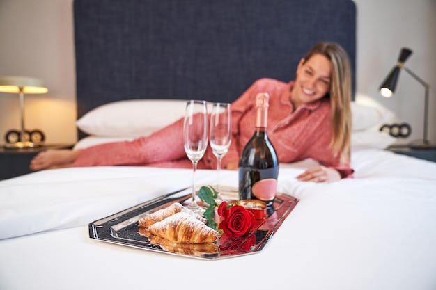 Happy female in sleepwear lying on bed near tray with glasses and bottle of champagne and red rose and delicious croissants presented for romantic occasion