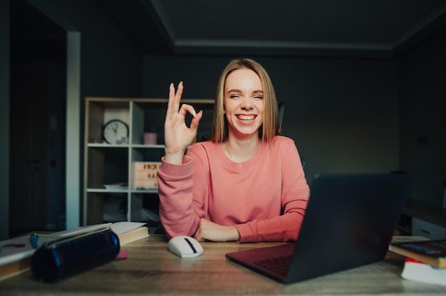 Foto felice studentessa seduta a casa alla scrivania con laptop e libri e in posa davanti alla telecamera