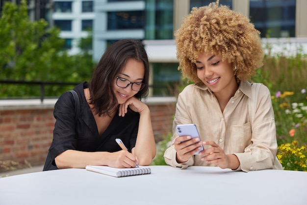 Happy female students meet together to write down some ideas\
for course work surf internet or browse social media on mobile\
phone plan organisation sits at table outdoors do university\
homework