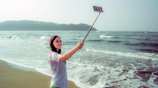 海の近くで自分撮りを撮る幸せな女性上からスマートフォンで一脚を使用してビーチで手を振っている海の近くで自分撮りを撮るうれしい女性