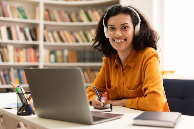 Happy female teacher in headphones using laptop and smiling to\
camera teaching online sitting in library indoors