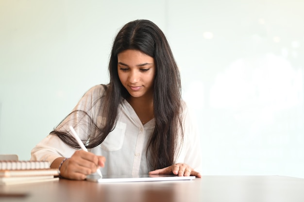 Happy female university student preparing for her exam on tablet while sitting on wooden desk.