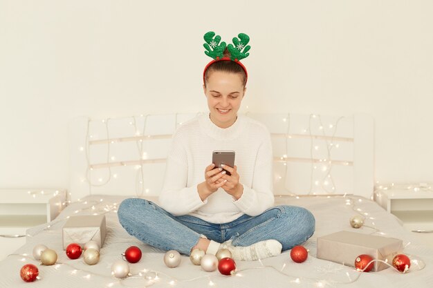 Felice femmina che indossa pullover e jeans in stile casual, in posa in camera da letto bianca, seduta sul letto, utilizzando lo smartphone per congratularsi online con capodanno e natale.