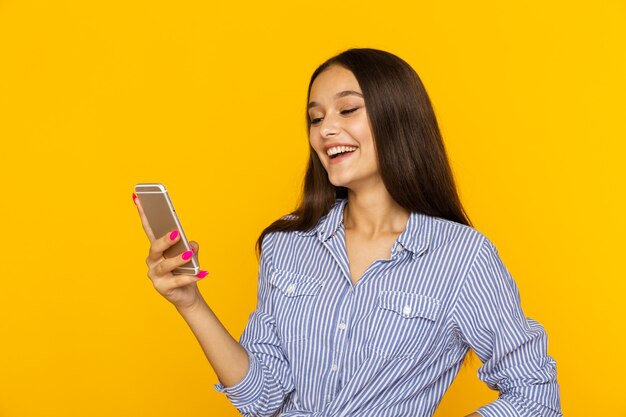 Happy female with mobile standing and smiling isolated over the yellow studio wall