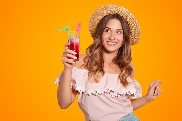 Happy female with positive smile, dressed in summer hat and blouse, holds summer cocktail, enjoys unforgettable rest, has positive expression