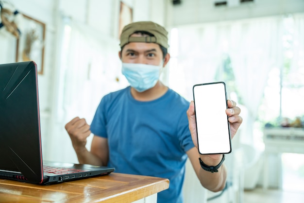 Happy freelance asian young man wearing face mask working with laptop computer and show mobile phone