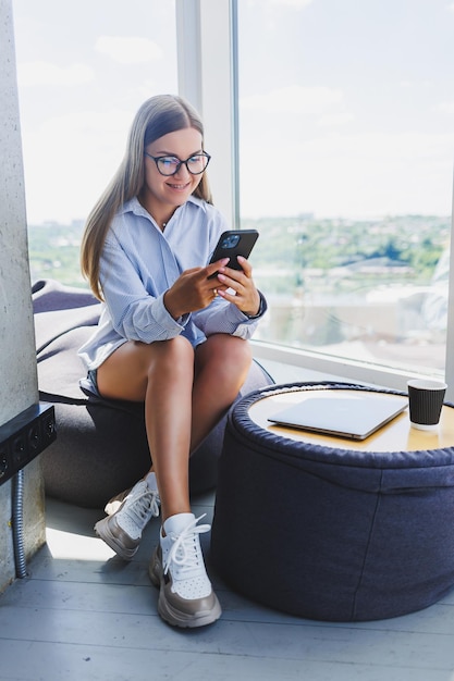Happy freelancer girl in classic glasses looking at phone and smiling while sitting in modern coworking space carefree millennial woman in glasses enjoying leisure time for communication