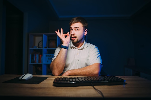 Happy freelancer man working on computer at night at home and\
showing ok gesture