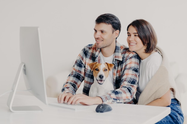 Happy friendly couple sit together with dog, make booking of\
hotel for future trip, keyboard and look at computer, chat online,\
pose at white desktop, connected to free wifi, browse internet\
together