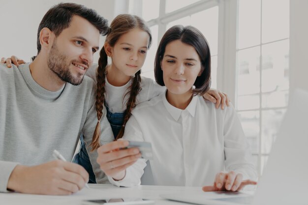 Happy friendly family of father mother and daughter sit in front of computer check balance on credit card make shopping online buy something necessary use high speed internet connection