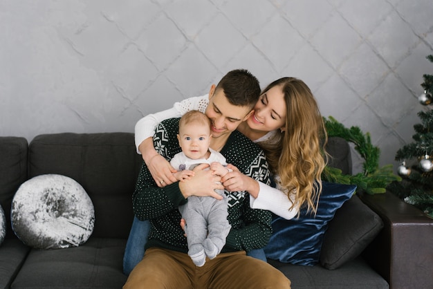 Happy friendly young family with a small child son sitting on the couch at the Christmas tree. They hug and smile