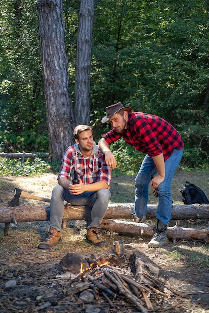 Happy friends on a camping trip relaxing by campfire group of\
friends enjoying picnic in the forest and drinking beer two\
lumberjack men making picnic tourists relaxing