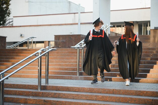 卒業式の日に幸せな友達 大学の建物の近くに立っている 2 人の陽気なうれしそうな学生の肖像画