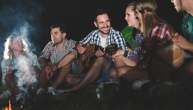 Happy friends playing music and enjoying bonfire in nature