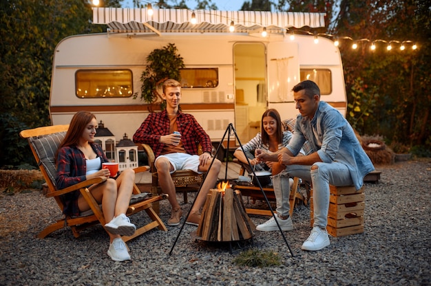 Amici felici che si siedono vicino al fuoco la sera, picnic al campeggio nella foresta