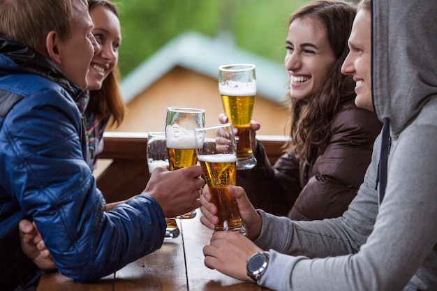 Счастливые друзья сидят с высокими бокалами пива в руке за деревянным столом