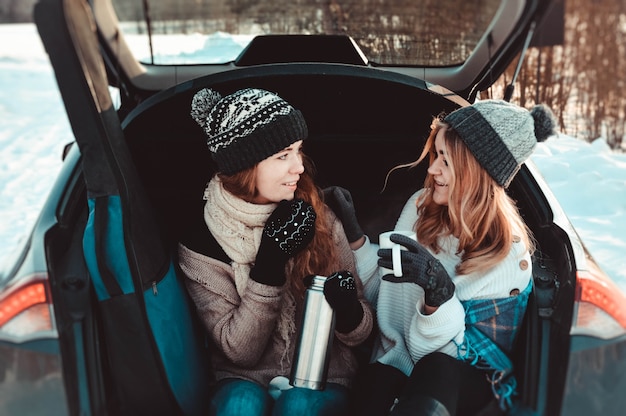 Happy friends in winter forest in the car. Two happy girls sit in the trunk of a car drinking coffee from a thermos, talking and laughing.