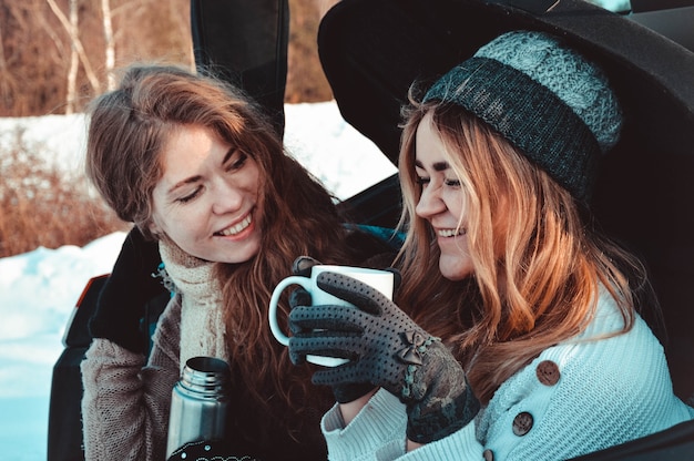 Happy friends in winter forest in the car. Two happy girls sit in the trunk of a car drinking coffee from a thermos, talking and laughing.