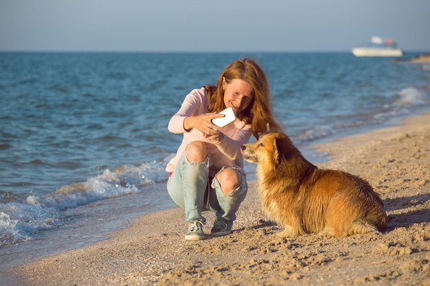 海のそばで幸せな楽しい週末-犬と一緒に笑顔の女の子がビーチでスマートフォンで自分撮りをします