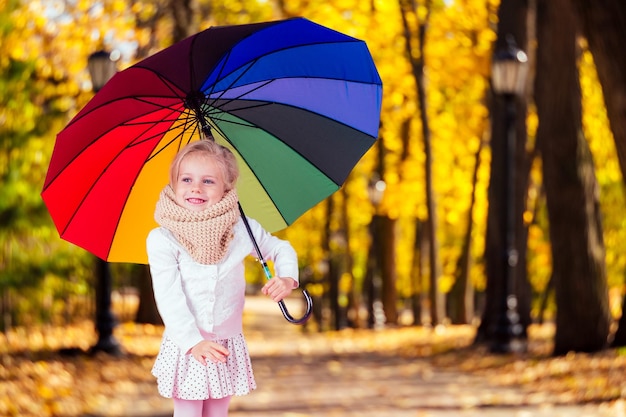 Happy funny beautiful blonde with blue eyes child with rainbow colorful umbrella in the autumn park Girl kid playing on the nature outdoors Family walk in the september forest yellow trees leaves