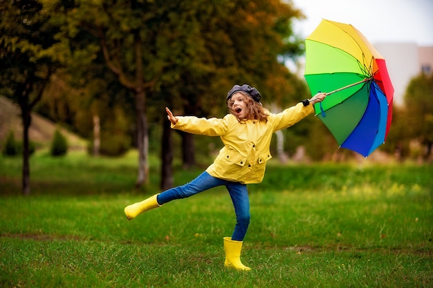Happy funny child girl with multicolor umbrella in rubber boots at autumn park.