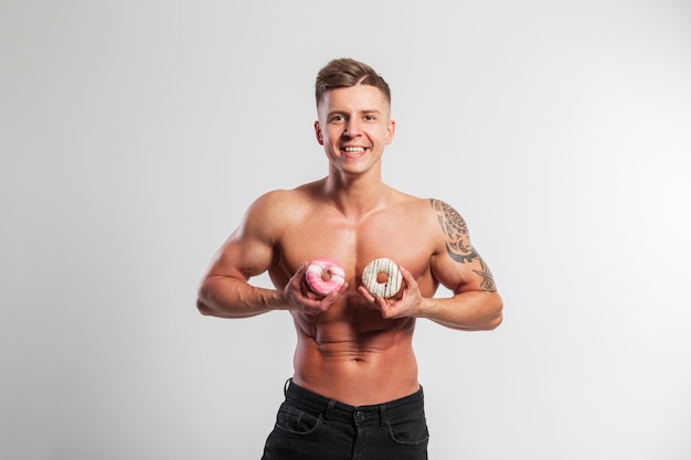 Happy funny fitness muscular man with naked torso holding donuts on white background Sports diet and sweets