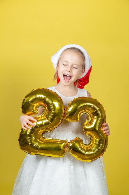 Ragazza divertente felice in cappello rosso della santa di natale con i numeri dell'aerostato su fondo giallo