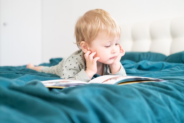 Счастливый смешной маленький мальчик в пижаме, читая книгу, лежа в кровати его родителей.