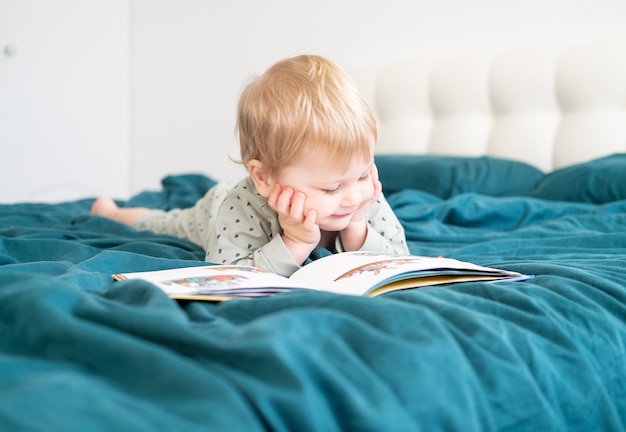 Photo happy funny little boy in pajamas reading book lying in his parents bed