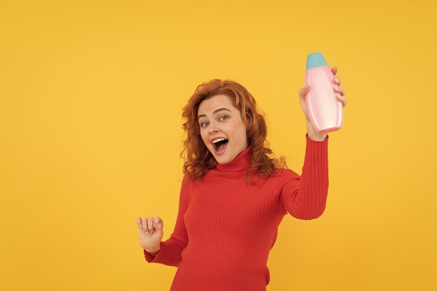 Felice ragazza rossa dai capelli ricci tinti che ballano presentando un prodotto per la cura del corpo di shampoo per la cura dei capelli