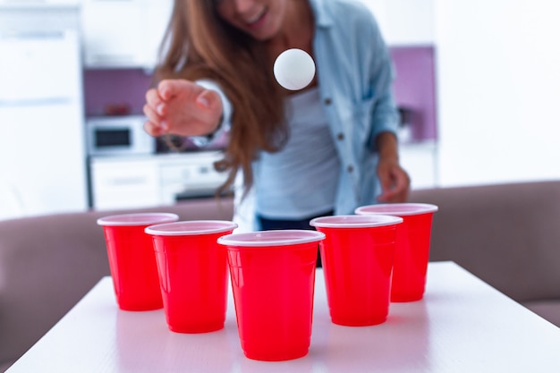 自宅のテーブルでビール卓球ゲームを楽しんでドリンクを飲みながら幸せな面白い女性